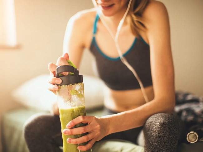 woman sitting on her bed with headphones on ready to sip on her pre-workout shake