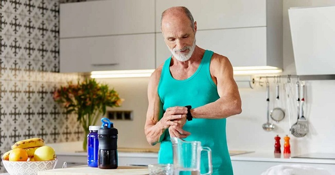 elderly man checking his watch standing behind a kitchen counter with a pre-workout shake in front of him